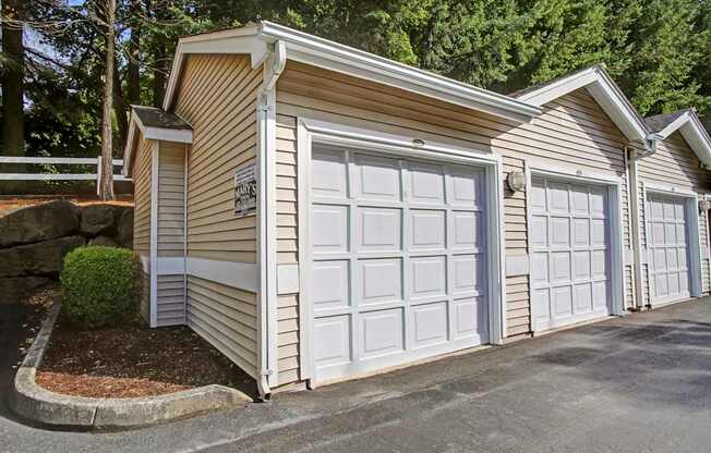 A detached garage building here at Aspen Creek showcasing a row of individual single-car garage doors with white paneled fronts, neutral horizontal siding, and pitched roofs, set along a paved drive and framed by mature evergreen trees, landscaped shrubs, and a quiet residential setting that offers convenient covered parking close to the community homes.