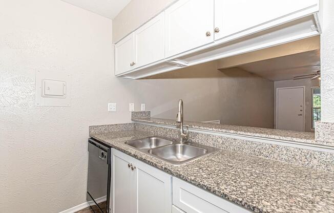 Modern kitchen featuring a granite countertop with double stainless steel sinks, white cabinetry, a silver dishwasher, and a neutral-colored wall. The kitchen has an open layout with a view into the adjoining living area.