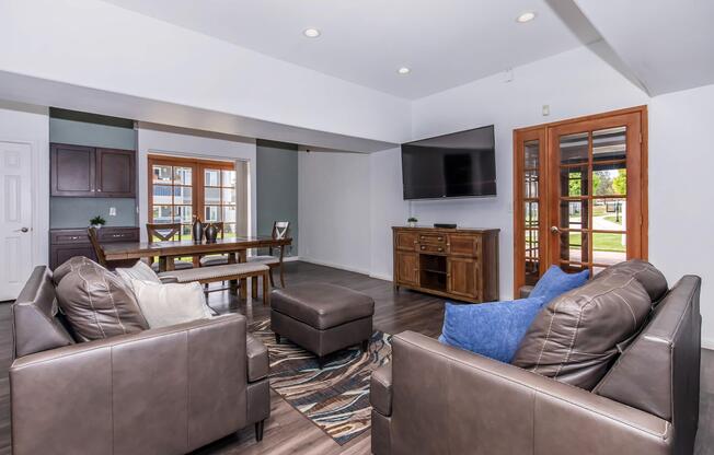 A modern living room featuring two brown leather chairs and a matching ottoman. In the background, there is a wooden dining table with chairs, a TV mounted on the wall, and large windows showing outdoor views. The floor is covered with a decorative area rug.