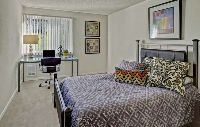 A comfortable bedroom here at Andorra featuring a neatly made bed with patterned bedding and accent pillows, a metal bed frame, and framed artwork on light-colored walls, complemented by soft carpeting, a window with vertical blinds, and a functional desk setup that creates a quiet space for rest and productivity.