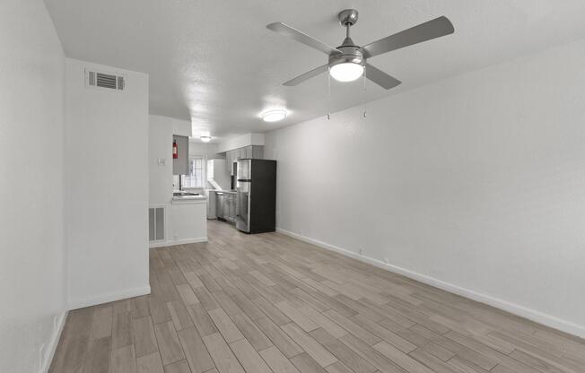 Interior view of a modern apartment featuring an open living space with light-colored wood flooring, a ceiling fan, and a window. The kitchen is visible in the background with stainless steel appliances and a bright ambience. The walls are painted white, contributing to a spacious feel.