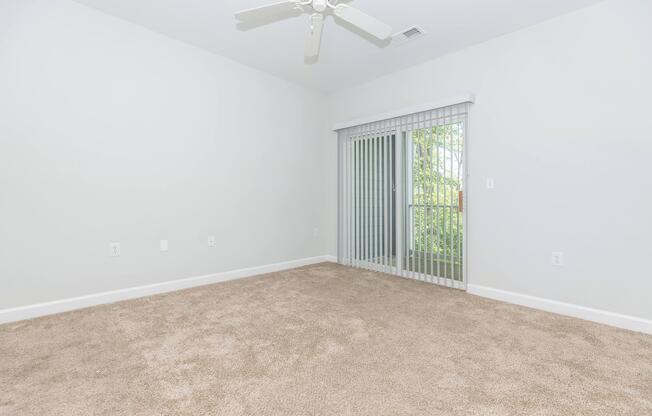 A spacious room with beige carpet and light gray walls. A ceiling fan is installed, and a large window with vertical blinds offers a view of greenery outside. The room is empty, providing a blank canvas for decoration or furniture placement. Natural light fills the space.