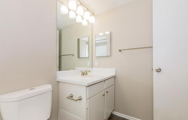 A modern bathroom featuring a white toilet and a light-colored countertop with a sink. Two mirrors are mounted above the countertop, and there are light fixtures above them. The walls are painted in a neutral tone, and a towel bar is visible beside the countertop. A door leads out of the space.