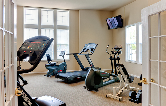 The fitness center, fitted with several fitness machines, carpet flooring, and a high ceiling.