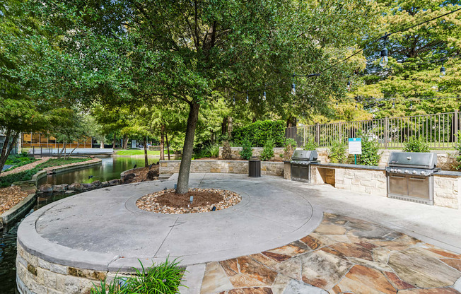 A tree in a circle of dirt with a stone wall and a grill in the background.