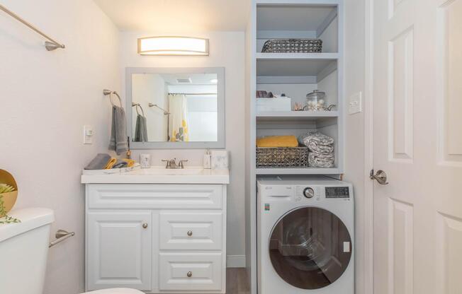 A modern bathroom featuring a white vanity with a sink and mirror, a washer and dryer unit next to it, and open shelves with neatly arranged towels and storage baskets. The walls are painted light colors, and there are decorative items on the counter. Natural light illuminates the space.