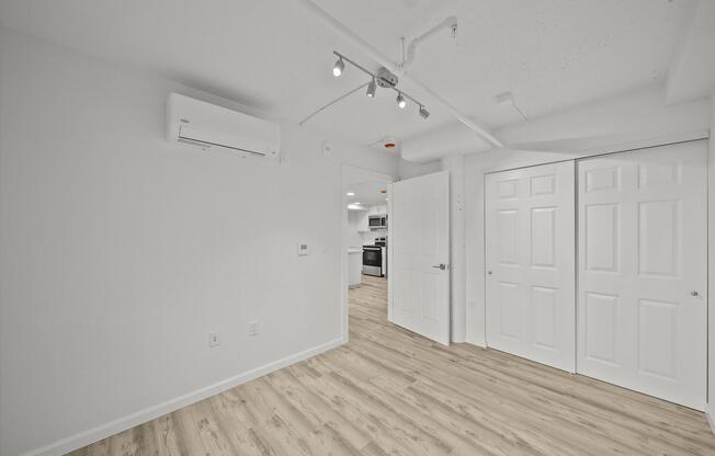 A well-lit, modern room featuring light-colored walls and wooden flooring. On the left, there is an air conditioning unit mounted on the wall, and on the right, a set of double doors leads to a closet. The room has recessed lighting and an open layout leading into a kitchen area visible in the background.