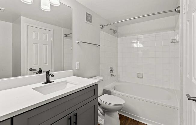 A modern bathroom featuring a white tiled bathtub and shower combination, a sleek white countertop with a black faucet, a toilet, and a mirrored vanity. The walls are painted in neutral tones, and there is a door leading out of the space, adding to the clean and contemporary design.