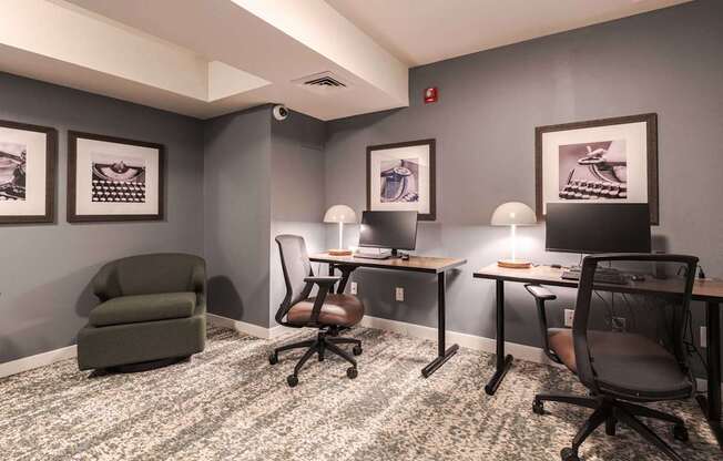 A room with grey walls and a carpeted floor with a green armchair, two desks with chairs, and framed pictures on the wall.