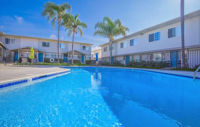 A sparkling blue swimming pool surrounded by palm trees, with two multi-story buildings in the background. Sunlight reflects off the water, creating a vibrant and inviting atmosphere. Poolside lounge chairs are visible, enhancing the resort-like feel of the scene.