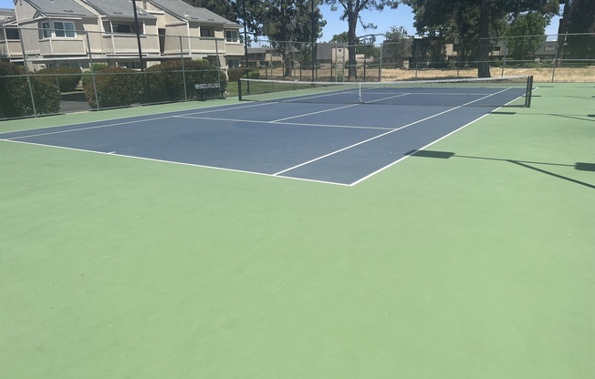 A tennis court with blue and green surfaces, featuring a net in the center. Surrounding the court are neatly trimmed hedges and residential buildings in the background, with clear skies overhead. The court appears well-maintained and ready for play.