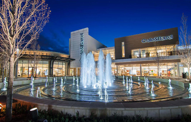 A Crate & Barrel store is lit up at night with a fountain in front.