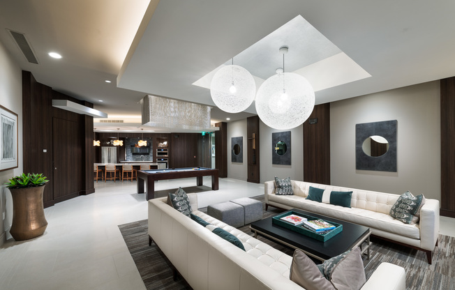 Clubhouse with dark wood-paneled walls, a pool table, two long white couches around a coffee table, and a resident catering kitchen with a wet bar.