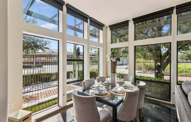 A dining table with chairs is set up in a sunny room with large windows.