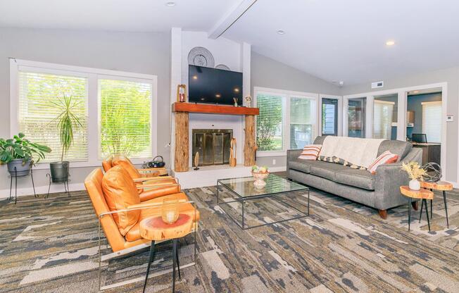 A modern living room featuring two stylish orange chairs and a gray sofa. A glass coffee table sits on a patterned rug, with a fireplace and a TV mounted on the wall. Large windows allow natural light, complemented by indoor plants and decorative items on the mantel and table.