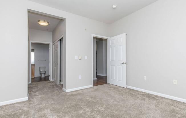 A spacious, empty room with light gray walls and carpeted floor. Two doorways lead to adjacent rooms, one showing a bathroom. A light fixture is visible on the ceiling, and the room has a clean, modern look with minimal furnishings.