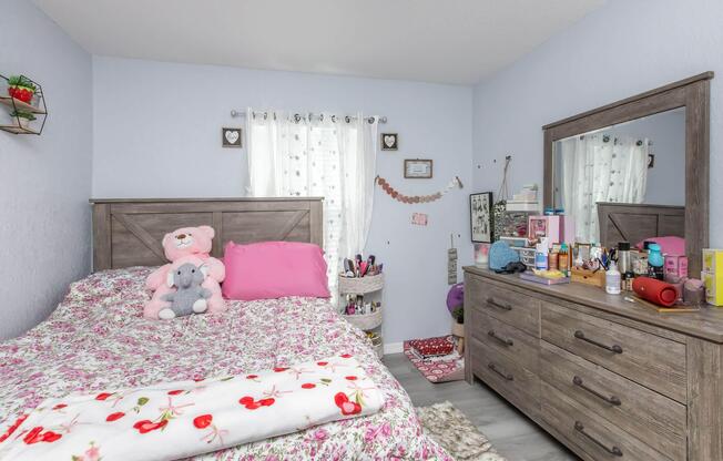 A cozy bedroom featuring a bed with floral bedding and a pink teddy bear, a wooden dresser adorned with beauty products, and a window with light curtains. Decorative items and personal touches create a warm atmosphere. The room is well-organized and inviting.