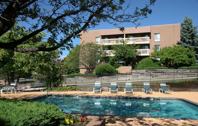 A sunlit outdoor pool area here at Eagle Ridge showcasing a clear swimming pool bordered by a spacious concrete deck with lounge chairs, mature trees providing shade, landscaped greenery and flowers in the foreground, and a multi-story brick apartment building with balconies rising in the background under a bright blue sky.