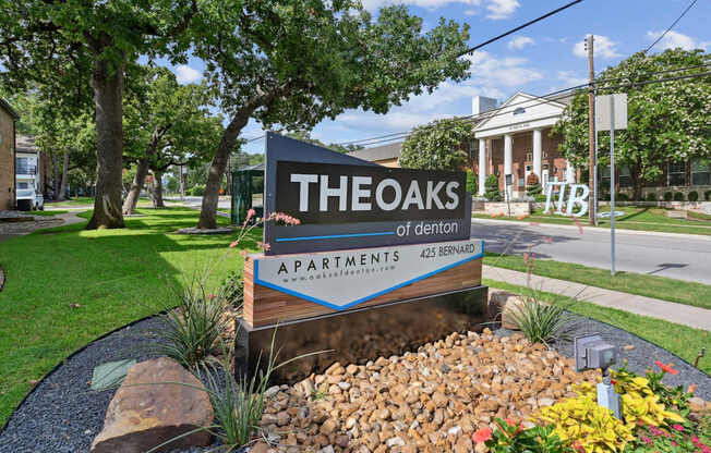 An entry sign for The Oaks of Denton Apartments is displayed in front of a landscaped area in Denton, TX