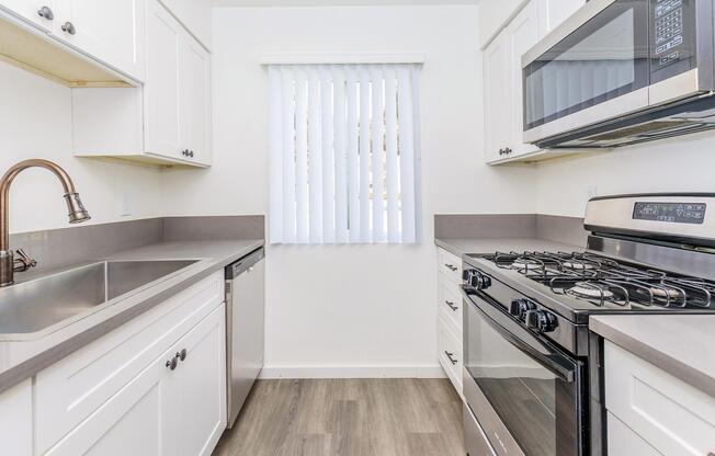 A modern kitchen featuring white cabinetry, a stainless steel sink, a dishwasher, a gas stove, and a microwave. The counter has a gray finish, and there are vertical blinds on the window, allowing natural light to enter the space. The flooring is light-colored laminate.