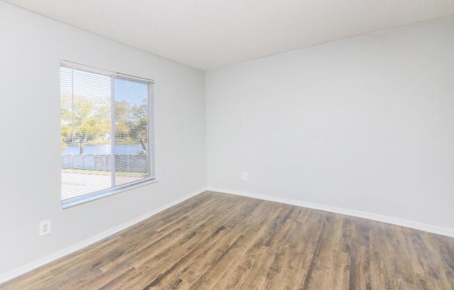A bright, empty room featuring light gray walls and a window with blinds, illuminating the space. The floor is covered with light wood-like laminate. Outside the window, trees are visible in soft focus, indicating a natural setting.