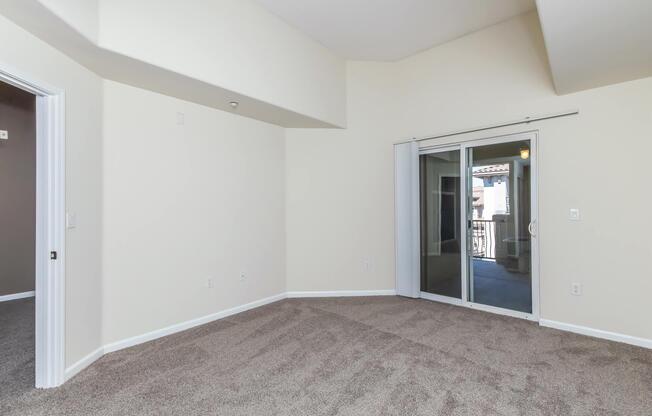 An empty room with light beige walls and carpeted flooring. It features a sliding glass door leading to a balcony, and a corner with a doorway visible. The ceiling is slightly angled, contributing to an open, airy feeling.