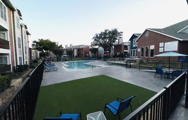 A residential complex featuring a swimming pool surrounded by lounge chairs. The area has artificial turf and is bordered by black fencing. Several trees are visible, along with apartment buildings in the background, creating a tranquil atmosphere.
