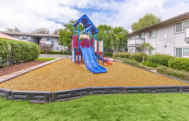 A colorful playground featuring blue slides and climbing structures, surrounded by a safety padding of wood chips. Lush green grass borders the play area, with several trees and buildings in the background under a partly cloudy sky.