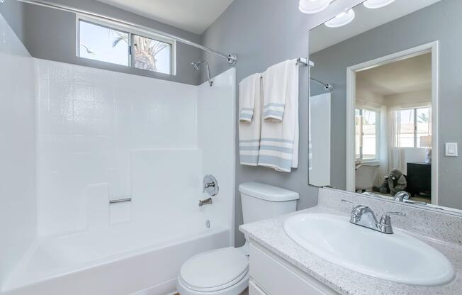 A modern bathroom featuring a white bathtub with a shower, light gray walls, a sink with a granite countertop, a toilet, and a large mirror. Towels hang on a rack, and natural light enters through a window, reflecting a cozy living space beyond.