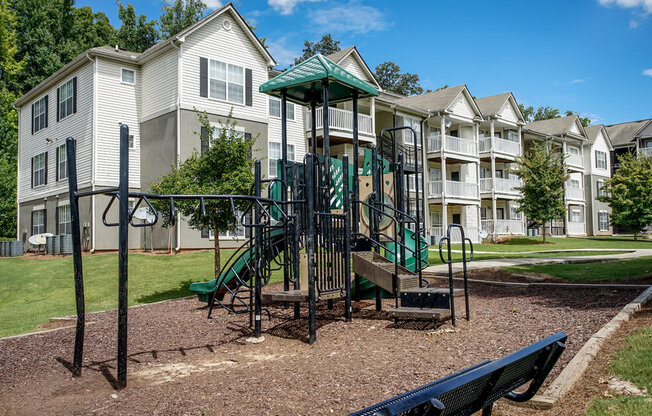 A playground with a green slide and a black bench in front of apartment buildings.