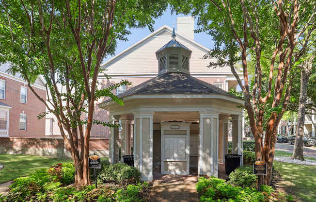 A small white building with a black roof and a bell tower is surrounded by trees and bushes.