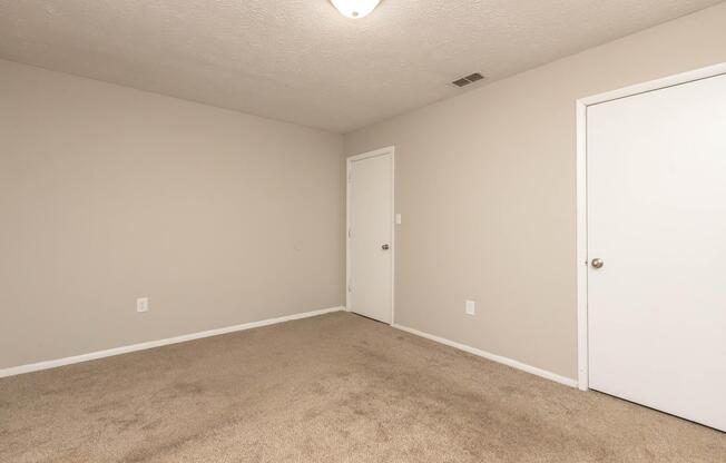 An empty room with beige walls and light brown carpet. There is a ceiling light fixture, a closed white door on the right, and a simple wall with no decorations or furniture. The overall space appears clean and spacious, ready for decoration or use.