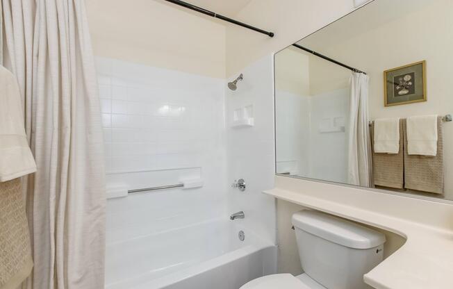 A clean bathroom featuring a bathtub with a shower, a white tiled wall, a large mirror, and a toilet. Light-colored towels are neatly hung on a rack, and a framed piece of artwork decorates the wall. Natural light enhances the bright and airy atmosphere of the space.