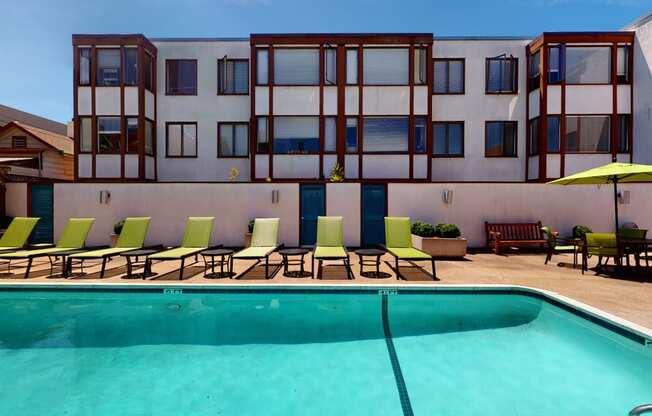 a swimming pool with green lounge chairs and a yellow umbrella in front of a white building