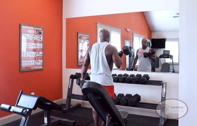 A man is weight lifting in front of a mirror inside of a fitness center with an orange accent wall.