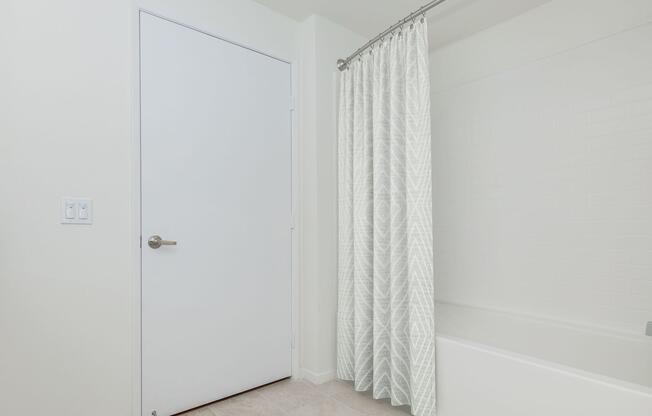 A clean, modern bathroom featuring a white door, a light-colored patterned shower curtain, and a bathtub with a tiled wall. The floor has light tiles and the walls are painted in a neutral tone, creating a bright and airy atmosphere.