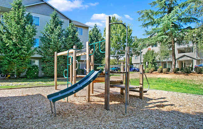 A neighborhood playground here at Allegro featuring a wooden play structure with climbing platforms, safety rails, and a curved slide set over a mulch play surface, surrounded by open grassy areas, mature trees, and nearby residential buildings, creating a family-friendly outdoor space under a clear blue sky.