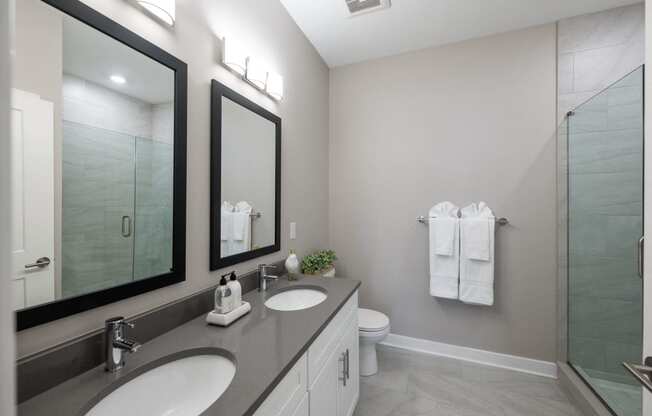 A bathroom with double vanity sinks, two large mirrors, a toilet, and a swinging glass door to a stand up shower with tile walls.