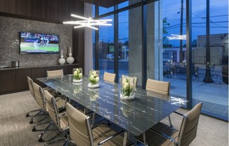Conference room equipped with 10-person conference table with rolling tan leather chairs, a modern angular light fixture, a full-width floor-to-ceiling window with curtains, and an HD presentation TV.