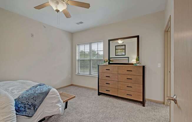 A bright bedroom here at Swan Creek with soft carpet, warm wood dresser, large window, and ceiling fan, creating a comfortable, inviting, and relaxing space filled with natural light.