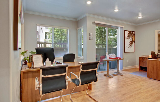 A leasing office here at Bridges with warm wood desks, black guest chairs, light wood floors, and bright windows with patio view, creating a clean, welcoming workspace.