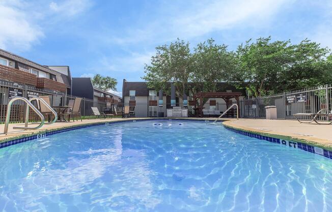 A clear swimming pool with steps leading in, surrounded by a patio area. In the background, there are several residential buildings and green trees under a blue sky. Sunlight reflects on the water's surface, creating an inviting atmosphere for relaxation and leisure.