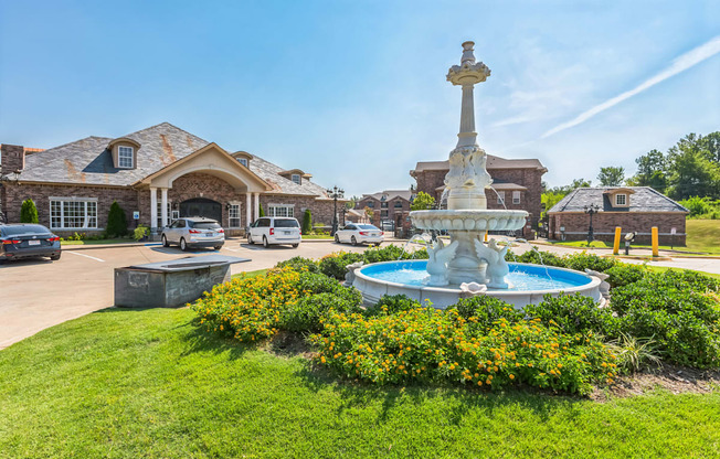 a fountain in front of a house with cars parked in a parking lot