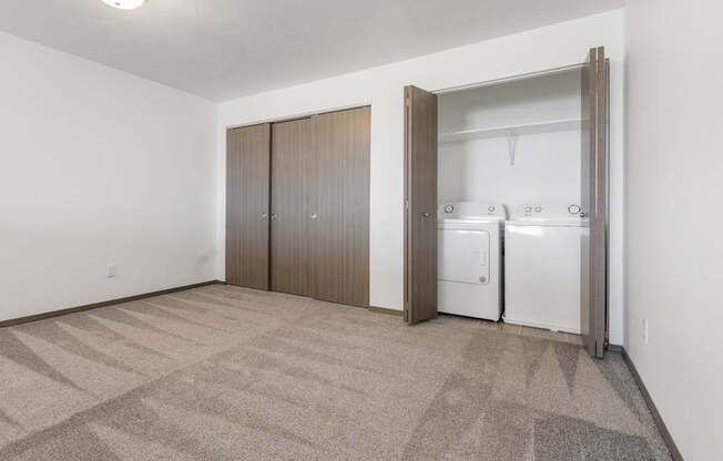 A room with a carpeted floor and a white wall with a fridge and cupboards in it.