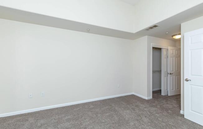 A spacious, empty room with beige walls and light brown carpet. There's a white closet with double doors on the right side. The ceiling is slightly elevated, and a light fixture is visible. The room appears bright and clean, suitable for various uses such as a bedroom or office.