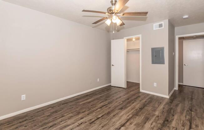 Empty room featuring light brown walls and flooring, a ceiling fan with light fixtures, a small closet with white doors, and a bathroom door on the right. The room has a neutral color palette and ample space, suitable for various furniture arrangements.