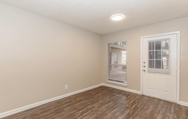 An empty room with light-colored walls and a textured ceiling. The floor is a dark, polished wood. There is a door on the right leading outside, and a window to the left with blinds, allowing natural light to enter. The space is uncluttered and ready for furniture.