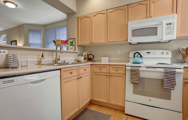 A welcoming kitchen here at Aspen Creek featuring light wood cabinetry with classic panel detailing, white countertops with a raised breakfast bar, and a full appliance suite including a range, microwave, dishwasher, and refrigerator, complemented by wood-style flooring, neutral walls, under-cabinet storage, and a pass-through opening that connects to the adjacent living area.