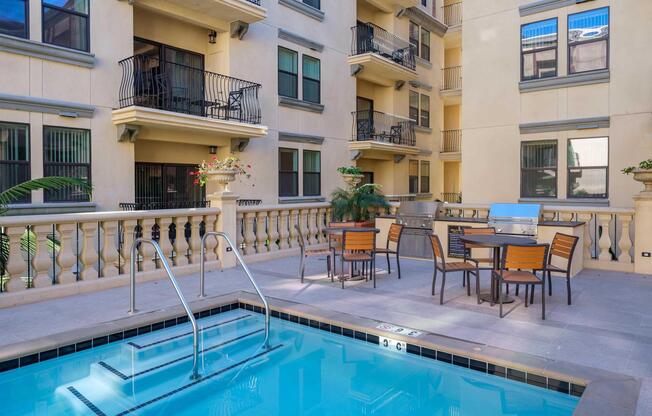 A serene apartment complex courtyard featuring a swimming pool with stairs, surrounded by a patio area with outdoor dining furniture and a grill. The building has multiple levels with balconies, and potted plants add a touch of greenery to the space.