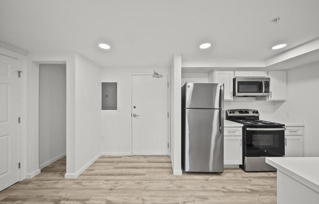 Modern kitchen and living area featuring a stainless steel refrigerator, microwave, and stove. The space has light-colored cabinetry and a neutral color scheme with a door leading outside. Natural wood flooring enhances the contemporary look.